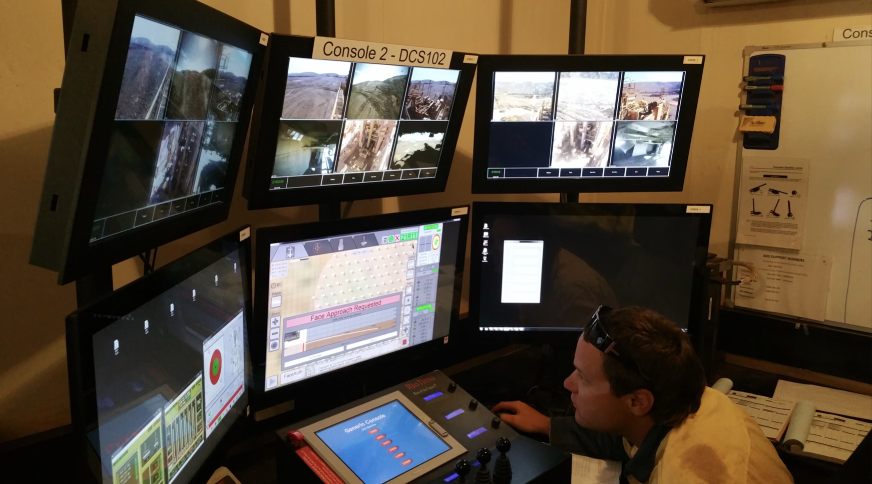 Drill operator monitoring six wall-mounted screens showing camera feeds and control software in a mine site control room